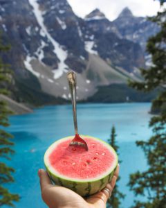 a hand holding a little watermelon with spoon at a mountain lake